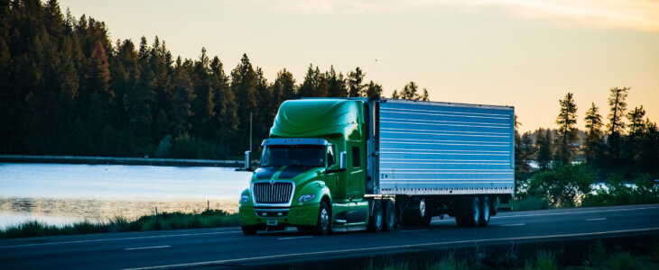 A green long-haul semi-truck with a refrigerated trailer, driving along a scenic section of interstate freeway just after sunrise, with trees and a lake in the background.